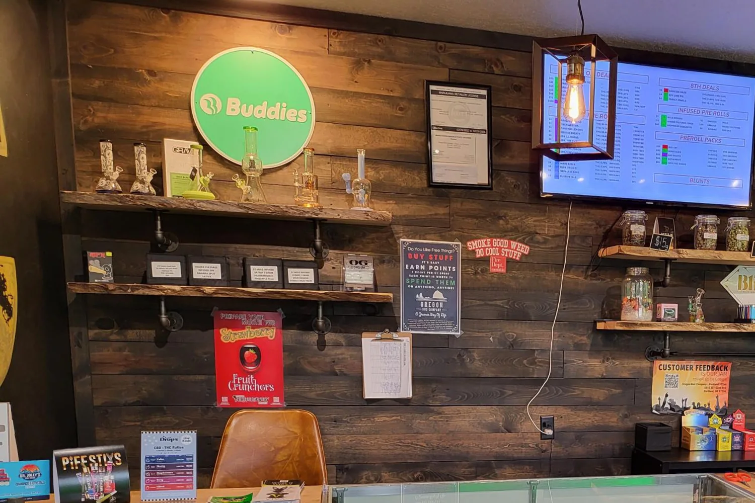 Wood-plank wall at Oregon Bud Company 122nd with glass pieces and flower jars on shelves, a menu screen and the glass counter in front
