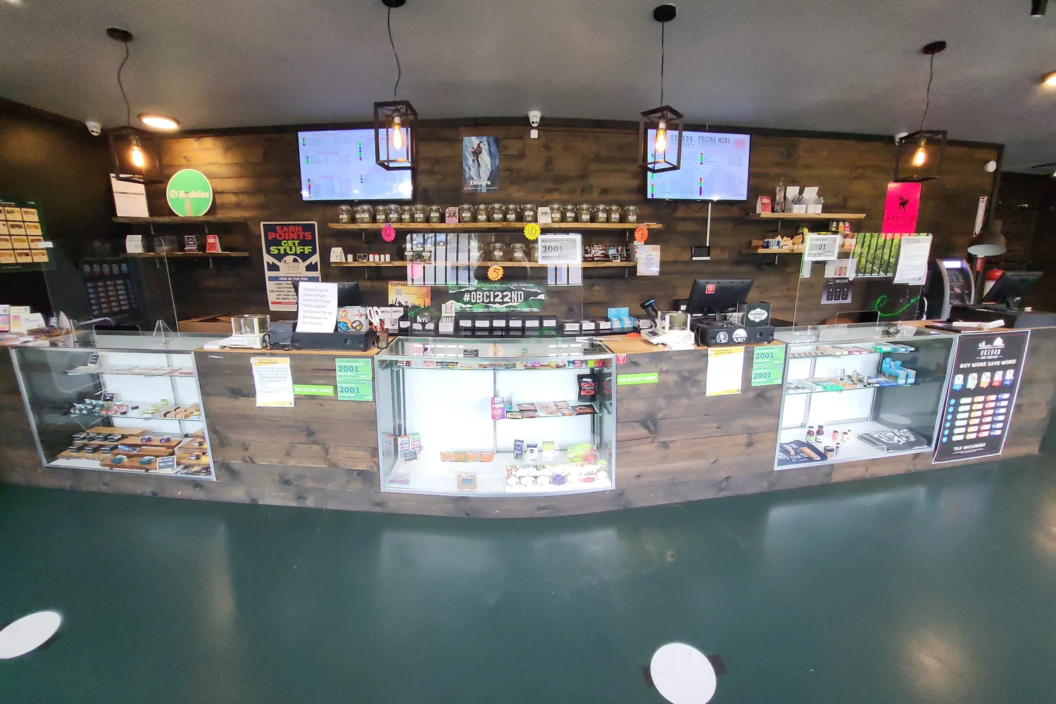 Long glass counter at Oregon Bud Company 122nd in Portland, with flower jars on wood shelves and two menu screens behind it